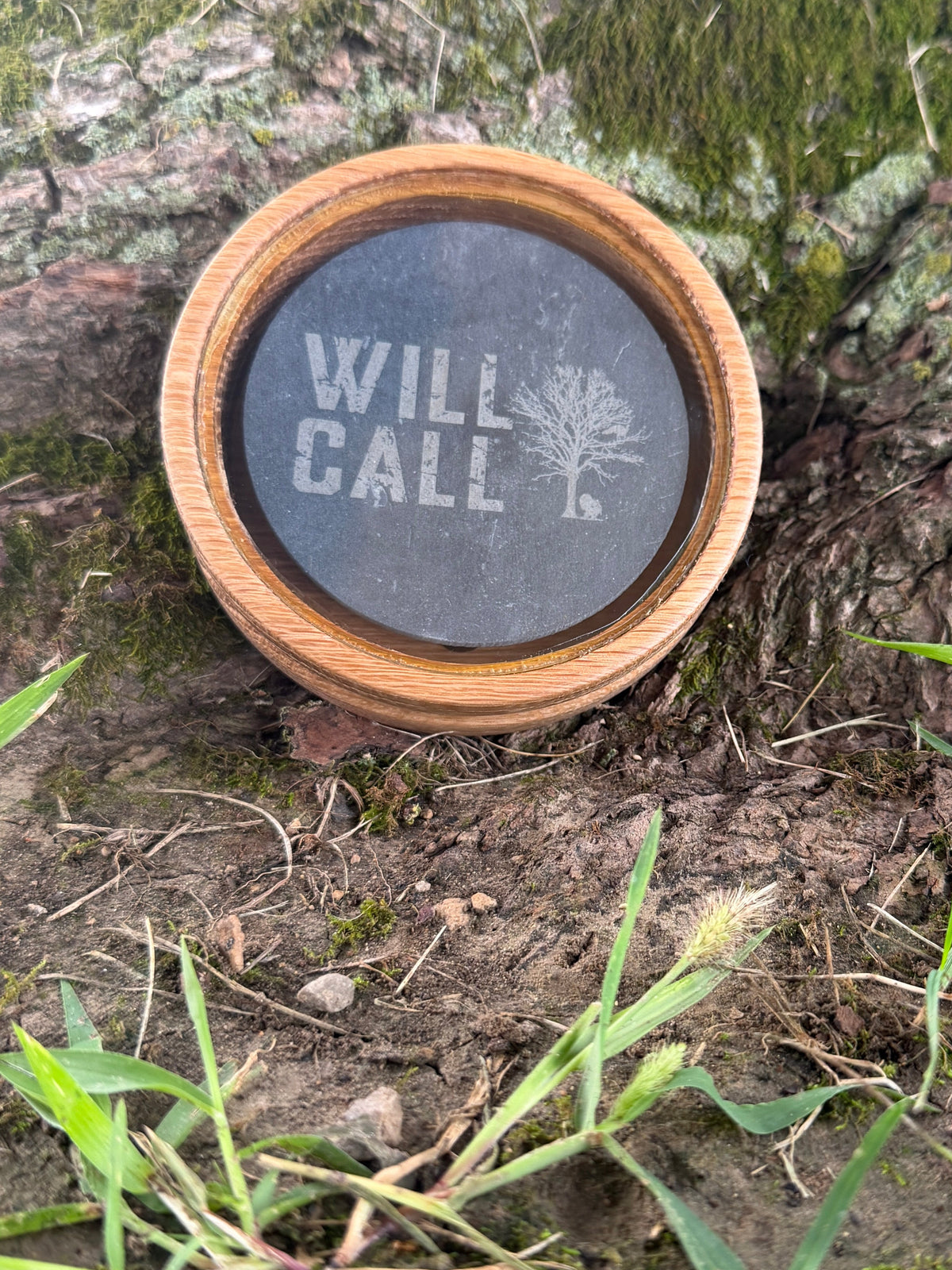 A round wooden-framed memorial clock or keepsake piece with a dark face and the words “WILL CALL” and a tree icon, placed outdoors on mossy tree bark with grass around it.
