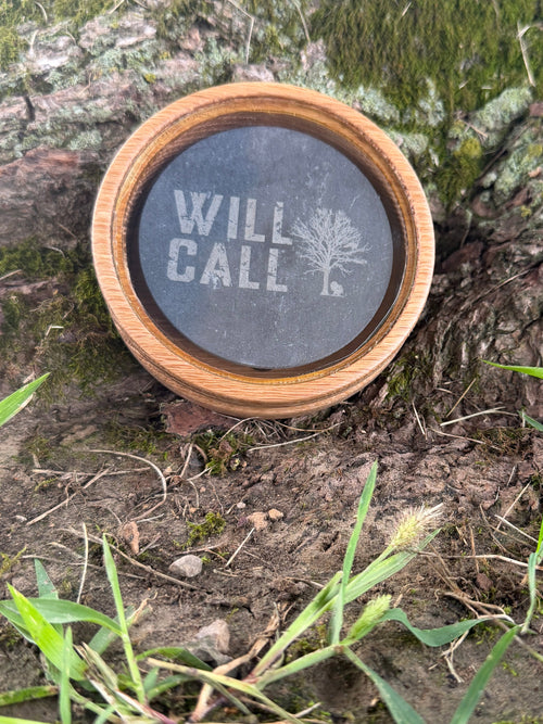 A round wooden-framed memorial clock or keepsake piece with a dark face and the words “WILL CALL” and a tree icon, placed outdoors on mossy tree bark with grass around it.