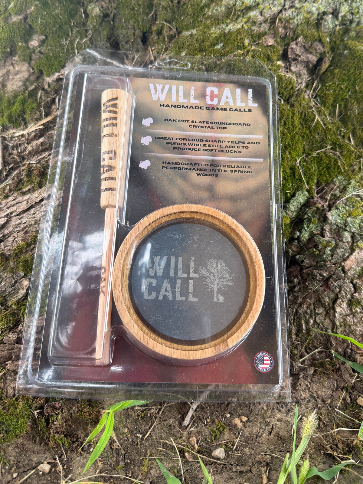 Packaged Will Call handmade game calls set, including a wooden call and a round black-and-wood holder, displayed on a mossy tree trunk outdoors.