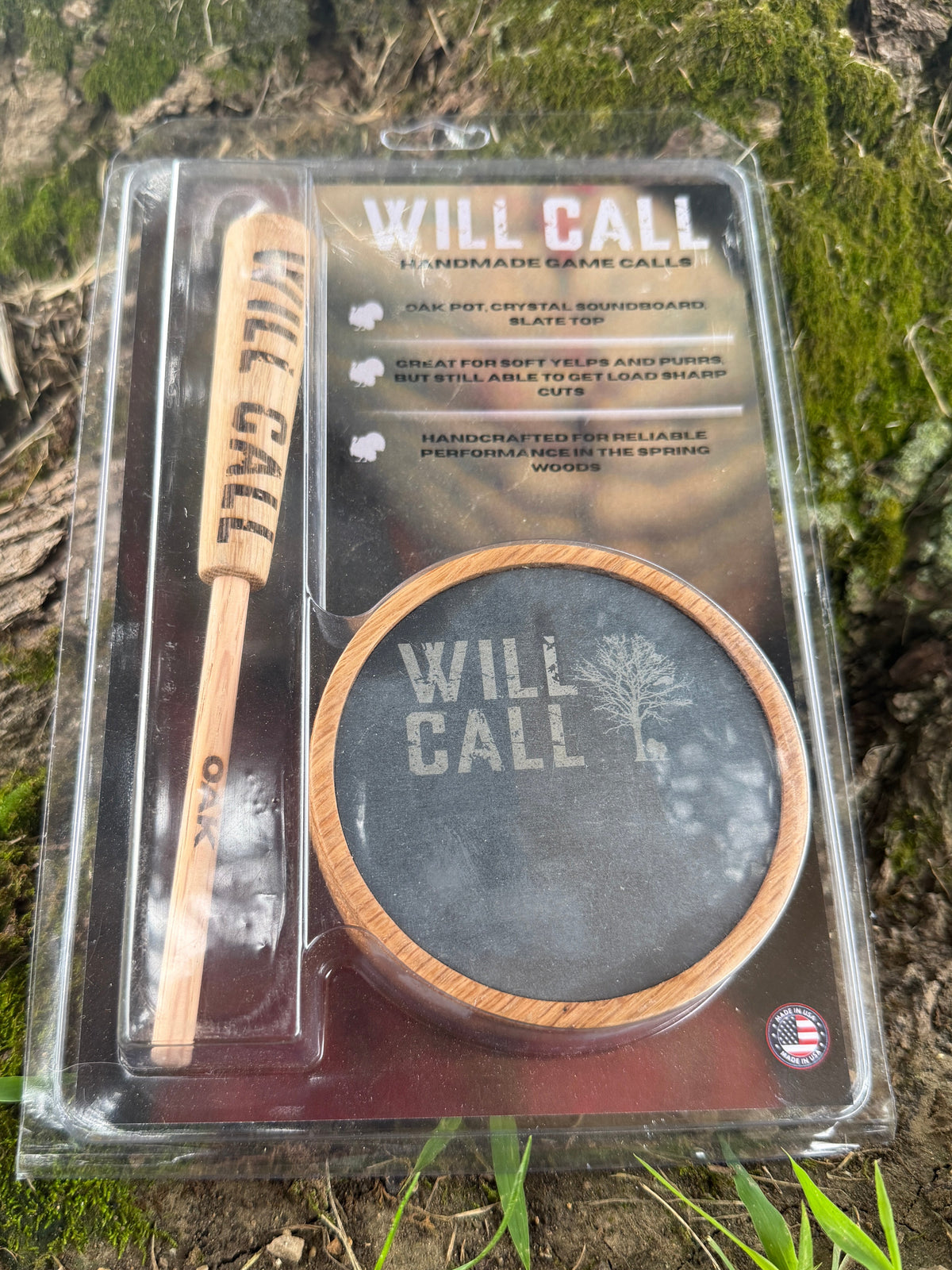 A packaged 'Will Call' Jamaican chain call set, containing a wooden stick tool and a round wooden tray, displayed in clear plastic packaging on mossy forest ground.