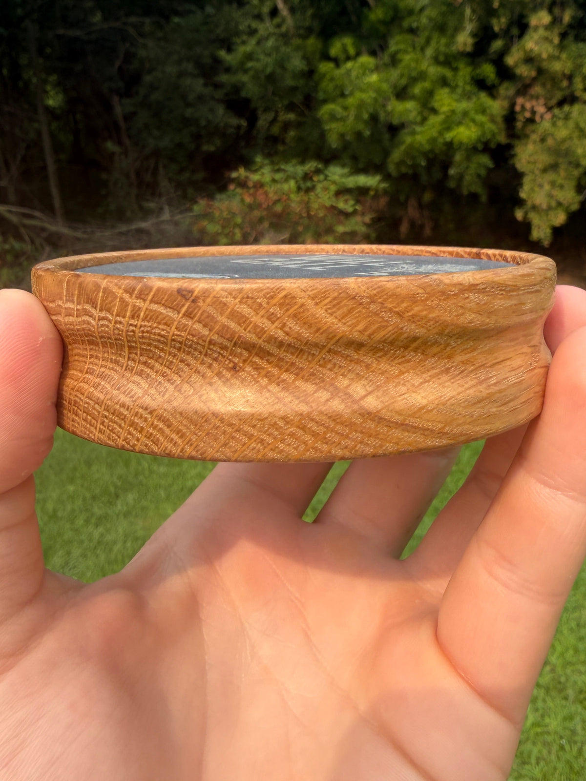 A hand holding a small, round, handcrafted wooden bowl with a visible wood grain pattern, set against a blurred background of green grass and trees.